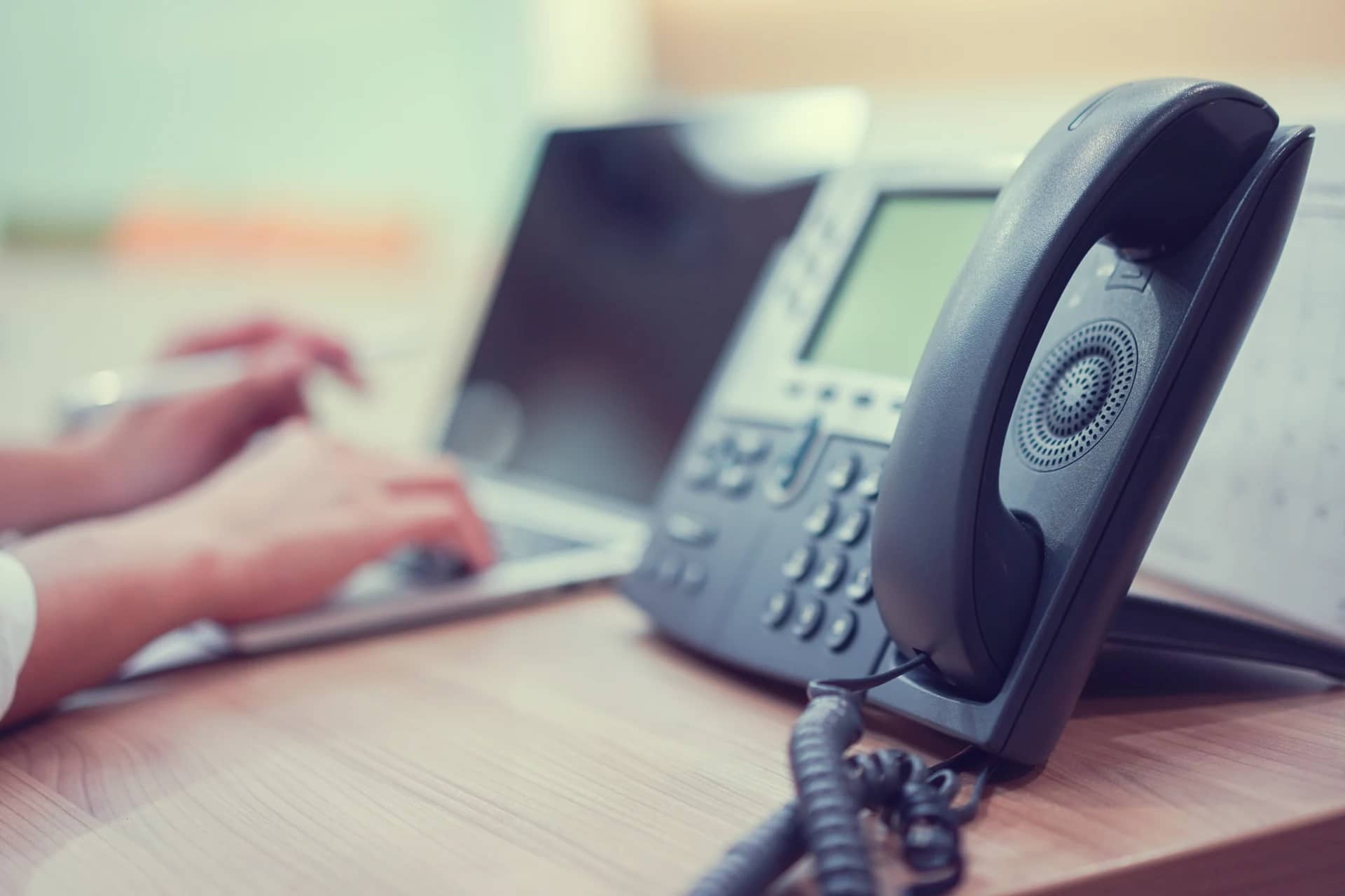 Téléphone de bureau au premier plan et mains qui tapent sur un ordinateur portable